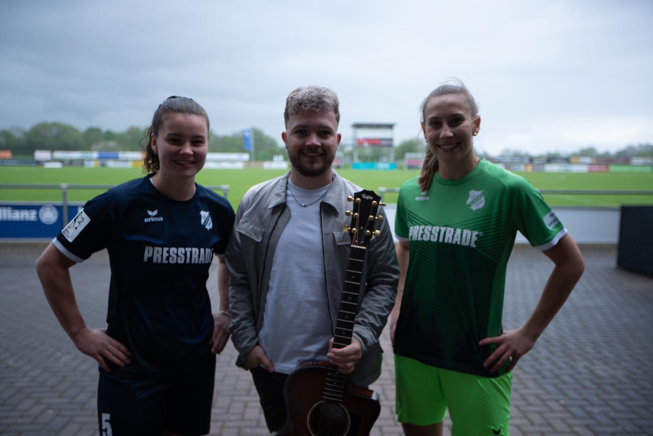 Links eine junge Frau mit Pferdeschwanz und dunklem Trikot mit dem Aufdruck "Presstrade", Mittig ein junger Mann mit T-Shirt, Jacke und Gitarre, rechts eine junge Frau mit Pferdeschwanz und grünem Trikot mit Aufdruck "Presstrade".