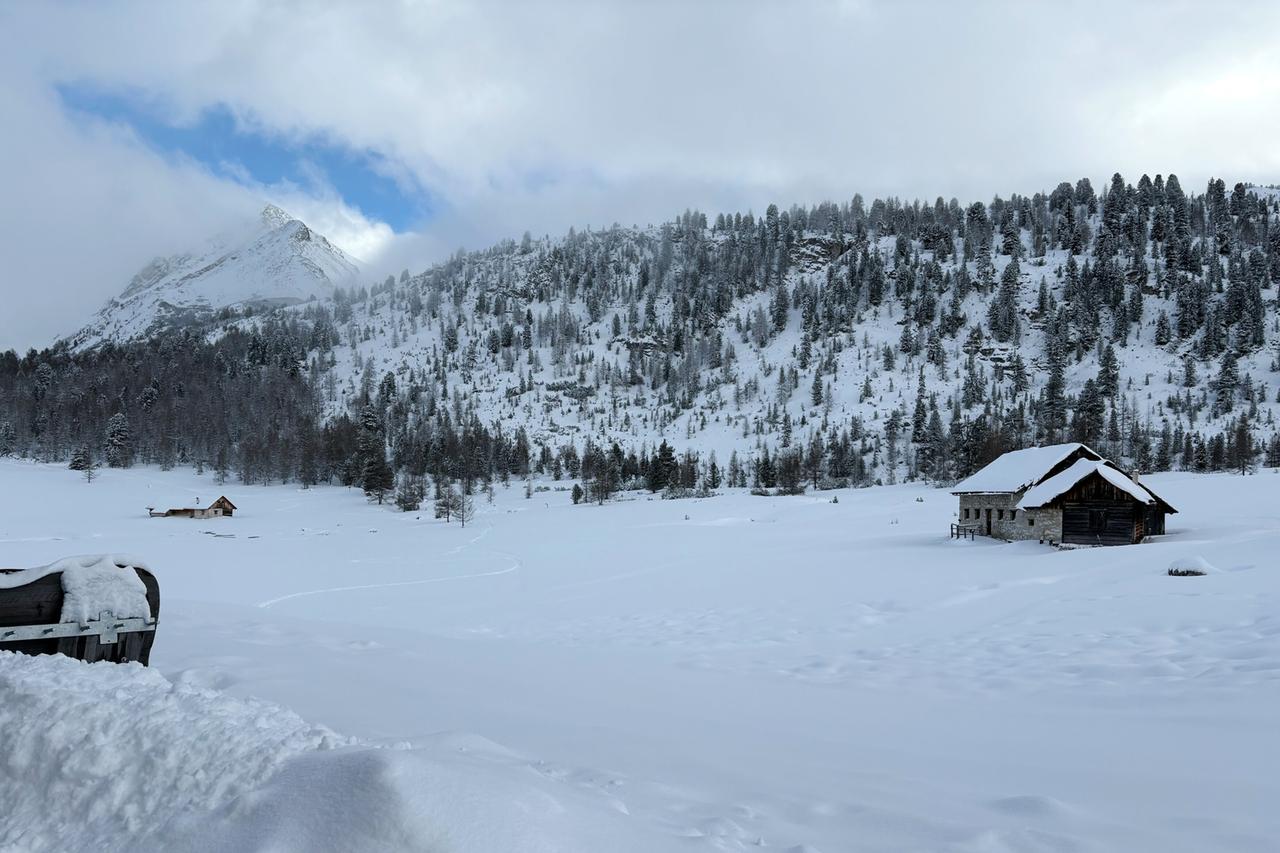 Ein verschneites Hochplateau unter blauem Himmel, eingerahmt von schroffen Dolomitengipfeln. Im Vordergrund eine Hütte mit Skispuren im Schnee.