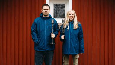 Ein Mann und eine Frau mit Mikrofonen vor einem roten Holzhaus.