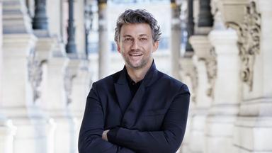  Der Tenor Jonas Kaufmann steht mit verschränkten Armen in einem prachtvollen Säulengang. Er ist komplett in Schwarz gekleidet.