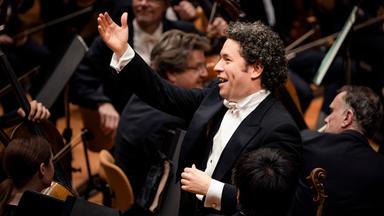 Der Dirigent Gustavo Dudamel steht vor dem Orchester. Er hebt die rechte Hand als Zeichen des Dankes an das Orchester. 