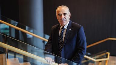 Ein Mann mit spärlichen grauen Haaren, gekleidet in einen dunkelblauen Anzug, steht auf einer Treppe.