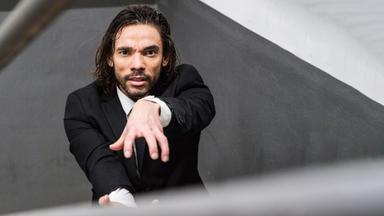 Blick von oben in ein Treppenhaus. Der Tänzer steht auf einer der Stufen und lehnt seinen Oberkörper nach hinten über das Metallgeländer. Bekleidet ist er mit einem schwarzen Anzug, einem weißen Hemd und einer schwarzen Krawatte.