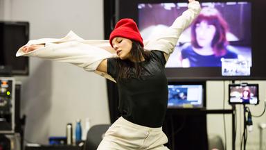 Die Tänzerin steht in einer Drehbewegung mit ausgestreckten Armen in einem Probenraum. Die Augen sind geschlossen. Im Hintergrund verschiedene Monitore. Sie trägt einen cremefarbenen Hosenanzug, ein schwarzes T-Shirt und eine rote Wollmütze.