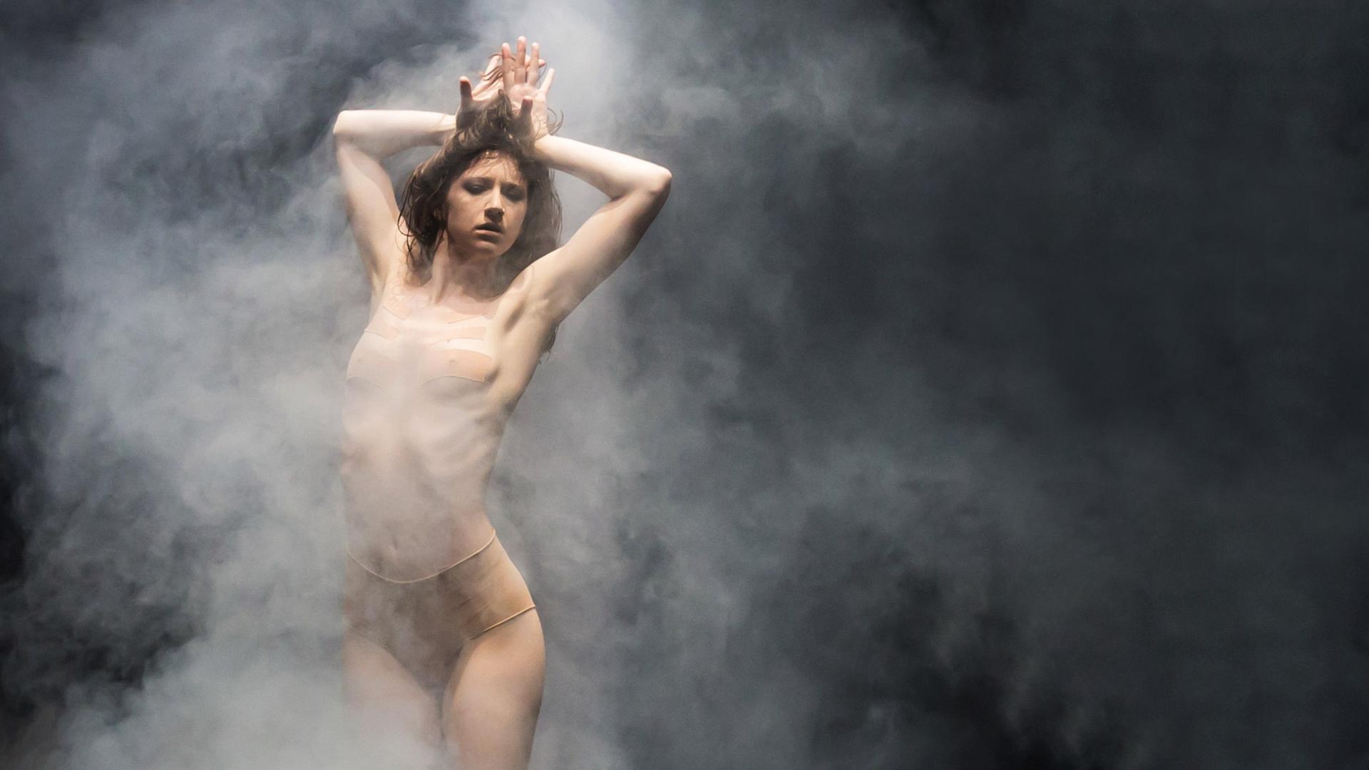 Die Tänzerin steht auf der Bühne, umgeben von Bühnennebel, die Arme verschränkt nach oben gestreckt. Bekleidet ist sie mit einem hautfarbenen Trikot.