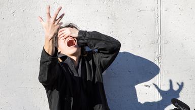 Choreografin und Tänzer haben die Arme verschränkt vor dem Gesicht. Eine Hand auf der Stirn, eine zur Seite. Sie blicken nach links.