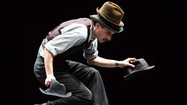 Der Tänzer Luca Pannacci läuft mit vorgebeugtem Körper und bebeugten Knien über die Bühne. Auf dem Kopf trägt er zwei Hüte übereinander. In jeder Hand hält er einen weiteren Hut.