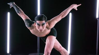 Der Tänzer steht auf der Bühne, ein Bein nach hinten gestreckt, das andere leicht angewinkelt nach vorne. Die Arme V-förmig nach oben gestreckt. Um ihn herum stehen Leuchtstoffröhren.