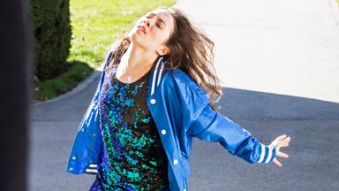 Die Tänzerin kniet im Probenraum auf dem Boden, den Oberkörper nach hinten gestreckt. Oben an der Decke hängt ein Bildschirm, über den die Choreografin Anweisungen erteilt.