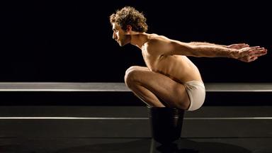 Der Tänzer sitzt in der Hocke auf dem Boden, die Arme nach vorne verschränkt.