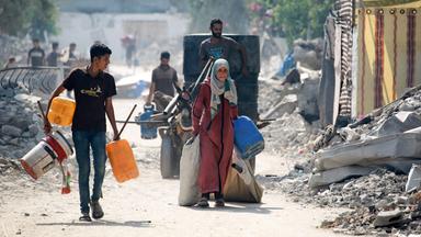 Menschen tragen Säcke und Kanister durch eine zerstörte Straße.