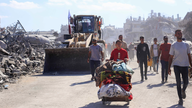 Menschen laufen durch eine zerstörte Stadt, Ein Junge schiebt einen Wagen mit seinen Habseligkeiten, am Straßenrand steht ein Bagger.