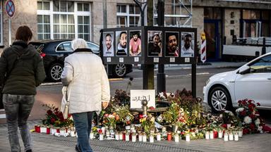 Auf einer Straße steht ein Metallgerüst mit Porträtfotos, davor stehen brennende Votivkerzen. Menschen gehen daran vorbei.