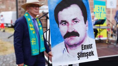 Vor einer Bühne hält eine Person ein großes Plakat mit dem Portätfoto eines Mannes mit Schnauzbart hoch.