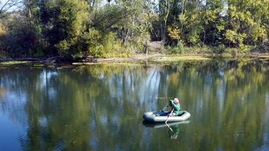 Das Bild zeigt eine ruhige Gewässerlandschaft mit einem Auwald im Hintergrund. Die Wasseroberfläche spiegelt die Bäume und das Ufer wider. In der Mitte des Bildes sitzt eine Person in einem aufblasbaren Boot, das grün ist, und hält eine Kamera in der Hand. Die Person trägt eine grüne Jacke mit roten Akzenten und hat weißes Haar. Es gibt Pflanzen und Bäume, die die Uferzone begrenzen. Die Stimmung ist friedlich und die Natur scheint üppig und lebendig zu sein. Diese Umgebung ist Teil einer dokumentarischen Ergründung über den Lebensraum von Tieren und Pflanzen in der Region.