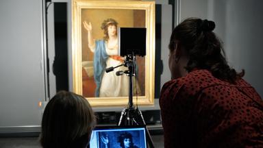 Ein Gemälde mit goldenen Rahmen zeigt eine Frau in Bewegung. Dieses wird beleuchtet und auf einem Laptop der mit der Lampe verbunden ist, wird ein Teil des Gemäldes noch einmal gezeigt. Zwei Frauen mit dem Rücken zur Kamera schauen sich die Aufnahme auf dem Laptop an.
