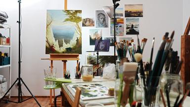 Auf einer Staffelei in einem Atelier ist ein Gemälde aufgestellt, auf dem eine Landschaft mit einer weißen Felsenbucht erkennbar ist.