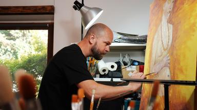 Ein Mann in schwarzem T-Shirt ist im Profil fotografiert, während er auf eine Leinwand malt. Darauf ist ein nackter, schwangerer Oberkörper erkennbar, vor einem einen gold-gelblichen Hintergrund.