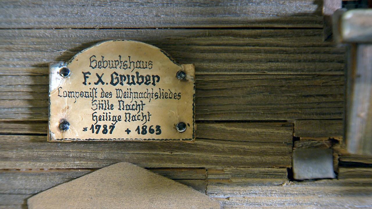 Das Bild zeigt eine metallene Tafel, die an einer Wand aus unbehandeltem Holz befestigt ist. Die Tafel trägt die Inschrift "Geburtshaus F. X. Gruber" sowie "Composer des Weihnachtsliedes Stille Nacht Heilige Nacht" und ist unterzeichnet mit den Jahren "1787" und "1863". Die Schrift ist in einer eleganten, geschwungenen Schriftart gehalten und wirkt leicht verblasst. Die Tafel hat eine rechteckige Form und ist mit zwei schwarzen Nägeln an der Wand befestigt. Die Holzoberfläche ist rau und zeigt sichtbare Maserungen sowie abgedunkelte Stellen, die auf ein gewisses Alter hinweisen. Das gesamte Motiv vermittelt einen historischen und kulturellen Kontext, der mit dem Komponisten Franz Xaver Gruber verbunden ist, der das bekannte Weihnachtslied "Stille Nacht" verfasst hat.