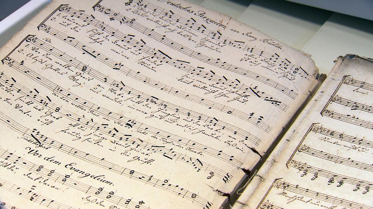 Das Bild zeigt alte Notenblätter, die sich auf einem Tisch befinden. Die Blätter sind vergilbt und zeigen handschriftliche Noten und Texte in einer alten, geschwungenen Schriftart. Die Noten sind in verschiedenen Zeilen angeordnet, und einige Zeilen sind durch zusätzliche Markierungen oder Striche ergänzt. Die Ränder der Blätter sind teils abgenutzt oder beschädigt. Der gesamte Eindruck vermittelt eine historische und nostalgische Atmosphäre, die auf die Bedeutung des Inhalts hinweist, möglicherweise im Zusammenhang mit dem berühmten Weihnachtslied "Stille Nacht."