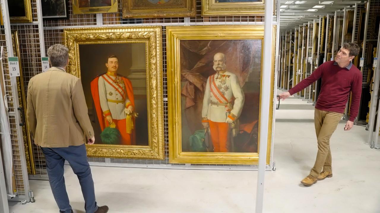 Zwei Männer betrachten Gemälde in einem Depot, darunter Porträts von Persönlichkeiten in militärischer Uniform.