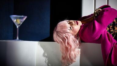 Das Bild zeigt eine weibliche Person mit langen, welligen, platinblonden Haaren, die in einer entspannten Position auf einer weißen Fläche liegt. Sie trägt einen auffälligen, pinken Mantel und darunter ein Kleid mit Leopardenmuster. Ihr Gesicht ist leicht zur Seite geneigt, und ihre Lippen sind leicht geöffnet, was einen nachdenklichen oder melancholischen Ausdruck vermittelt.  Neben ihr steht ein Glas mit einem Drink, in dem sich eine Zitronenscheibe befindet. Der Hintergrund ist dunkel, was einen interessanten Kontrast zur hellen Kleidung der Person bietet. Der Lichteinfall schafft Schatten, insbesondere um das Gesicht und die Haare der abgebildeten Person, und verstärkt den dramatischen Eindruck der Szene.