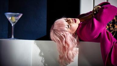 Das Bild zeigt eine weibliche Person mit langen, welligen, platinblonden Haaren, die in einer entspannten Position auf einer weißen Fläche liegt. Sie trägt einen auffälligen, pinken Mantel und darunter ein Kleid mit Leopardenmuster. Ihr Gesicht ist leicht zur Seite geneigt, und ihre Lippen sind leicht geöffnet, was einen nachdenklichen oder melancholischen Ausdruck vermittelt.  Neben ihr steht ein Glas mit einem Drink, in dem sich eine Zitronenscheibe befindet. Der Hintergrund ist dunkel, was einen interessanten Kontrast zur hellen Kleidung der Person bietet. Der Lichteinfall schafft Schatten, insbesondere um das Gesicht und die Haare der abgebildeten Person, und verstärkt den dramatischen Eindruck der Szene.