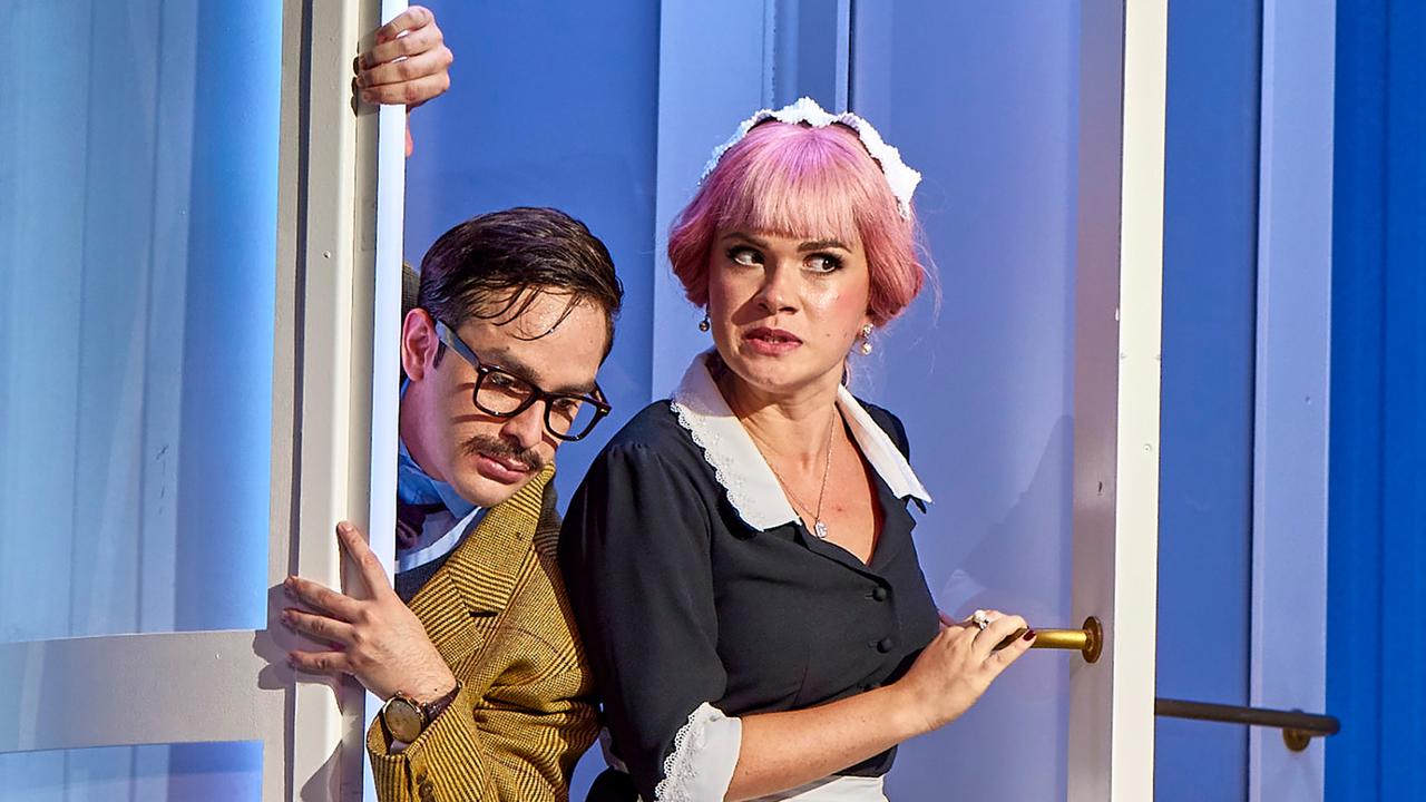 Das Bild zeigt zwei Charaktere aus der Kammeroper „Gelegenheit macht Diebe“ von Gioachino Rossini. Auf der linken Seite steht ein Mann mit einer Brille, der ein kariertes Jackett und ein Hemd mit einer Krawatte trägt. Er schaut nervös nach hinten und stützt sich an einer halb geöffneten Tür ab.  Rechts von ihm steht eine Frau mit einem Kleid in Schwarz und einem weißen, schulterfreien Kragen. Ihr Haar ist rosa und sie trägt eine weiße Haube. Ihr Gesichtsausdruck wirkt überrascht und angespannt. Die beiden scheinen sich miteinander zu unterhalten oder sind in ein heimliches Gespräch verwickelt.  Der Hintergrund des Bildes ist hell erleuchtet, überwiegend in blauen Tönen, was eine theaterähnliche Atmosphäre schafft. Es gibt auch eine zweite Tür hinter ihnen, die zum Raum gehört. Die Szenerie vermittelt eine humorvolle und spannende Stimmung, passend zur Handlung der Oper.