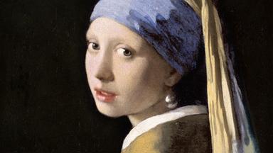 Porträt einer jungen Frau mit blauem und gelbem Kopftuch, gelber Kleidung und einem großen Perlenohrring vor dunklem Hintergrund.