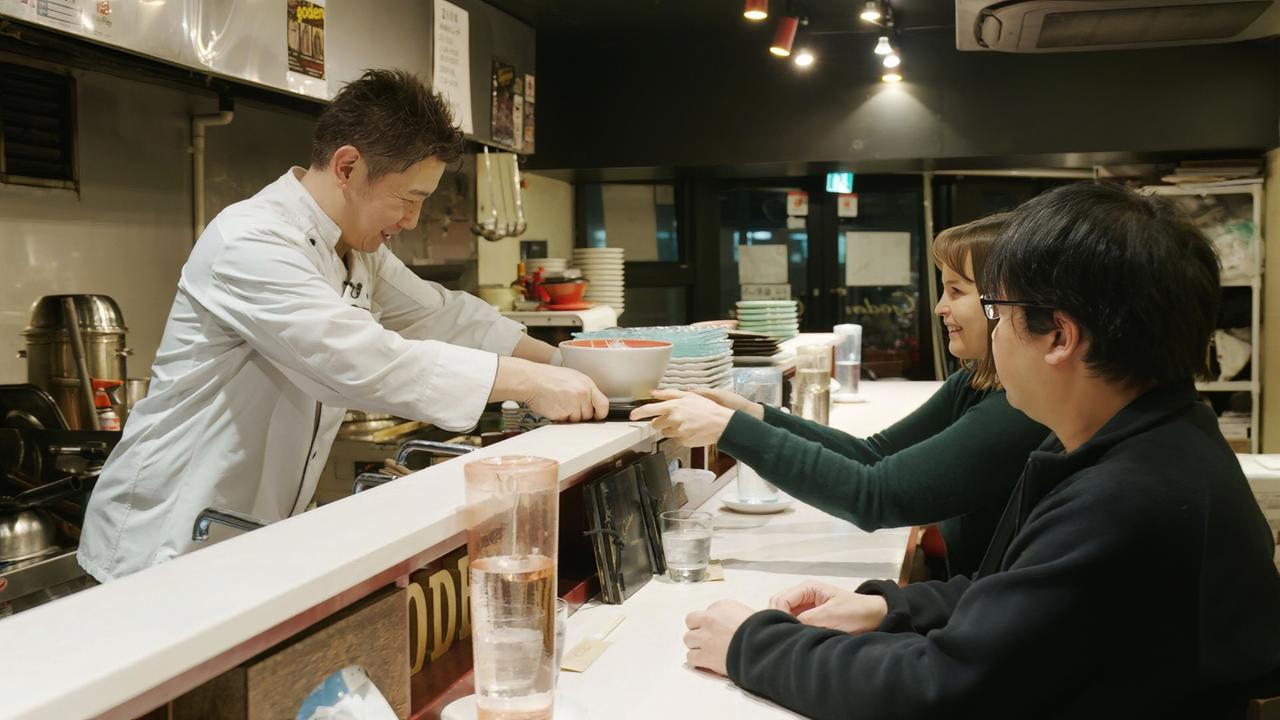 Eine Reporterin sitzt mit einem Mann an einer Theke in einem japanischen Restaurant, während ein Koch ihr eine frisch zubereitete Schüssel mit Essen überreicht.