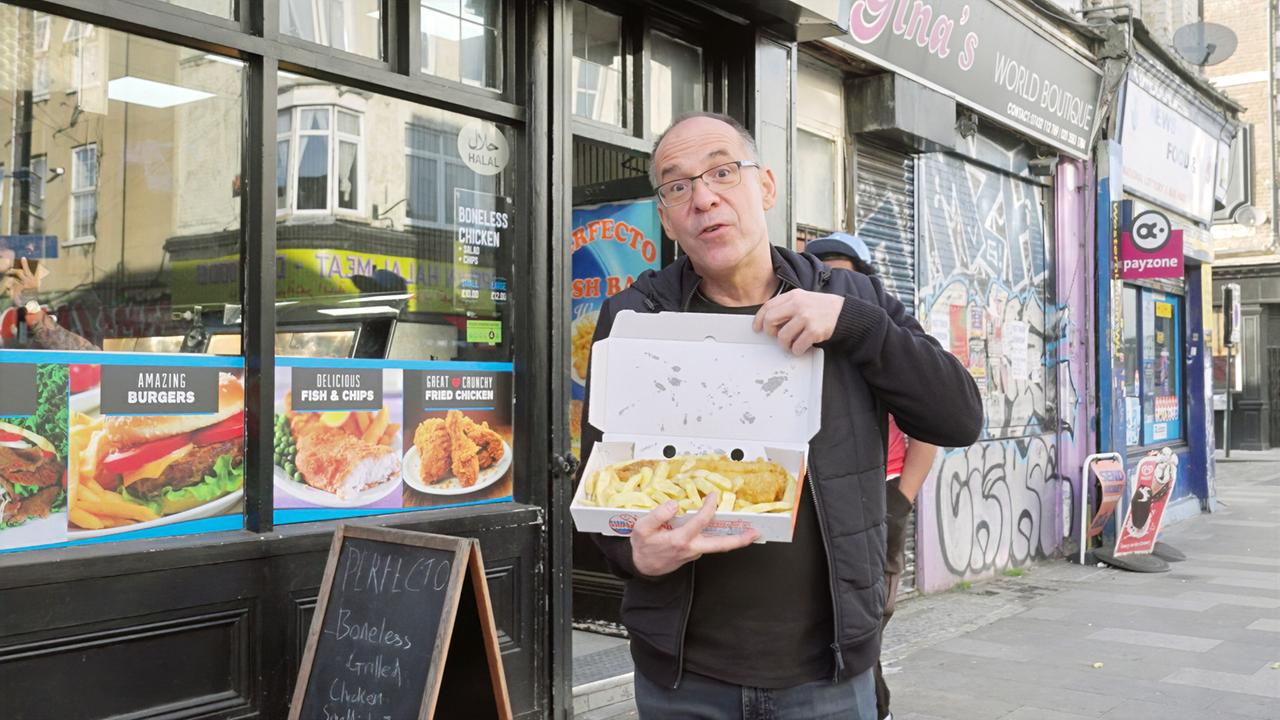 Ein Mann steht vor einem Imbiss in einer Londoner Straße und hält eine geöffnete Box mit Fish and Chips in der Hand. Im Hintergrund sind Graffiti und Geschäftsschilder zu sehen.