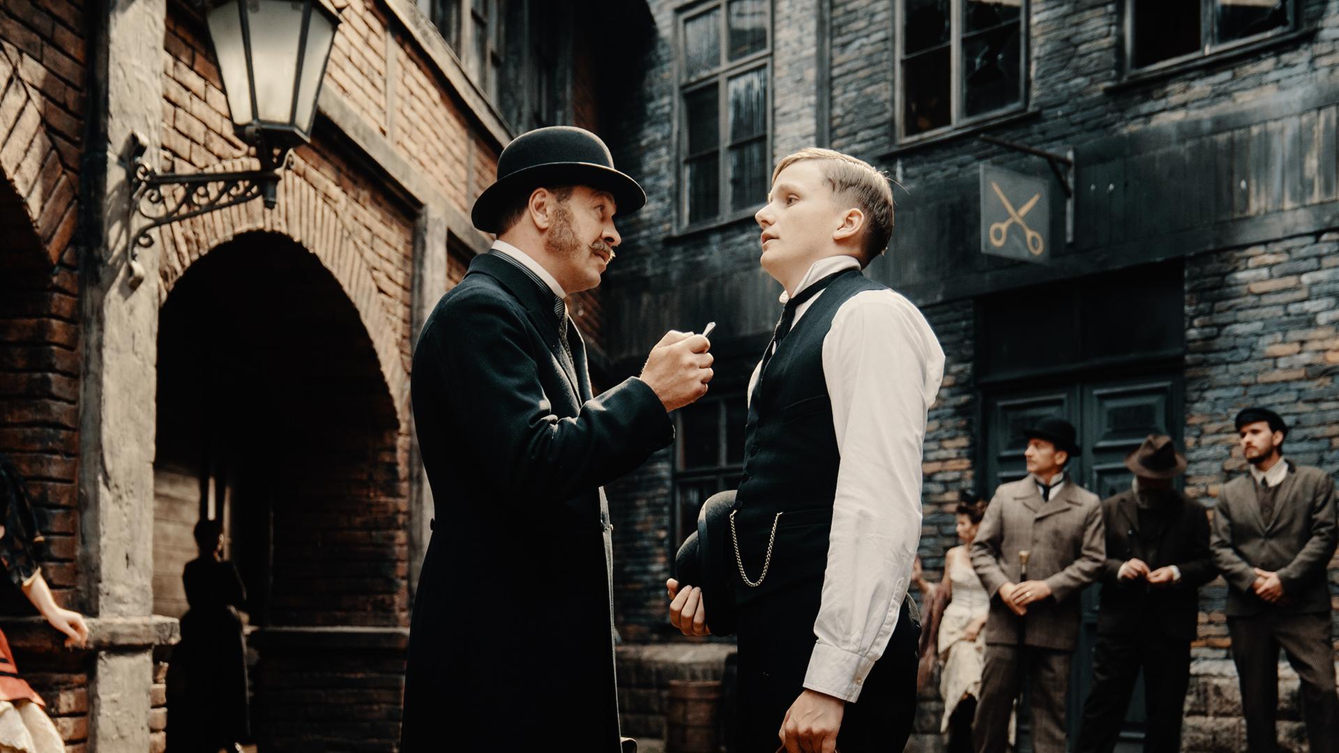 Nachgestellte historische Szene: Zwei Männer in historischer Kleidung stehen auf einer Straße und schauen sich kritisch an. Im Hintergrund stehen weitere Männer.