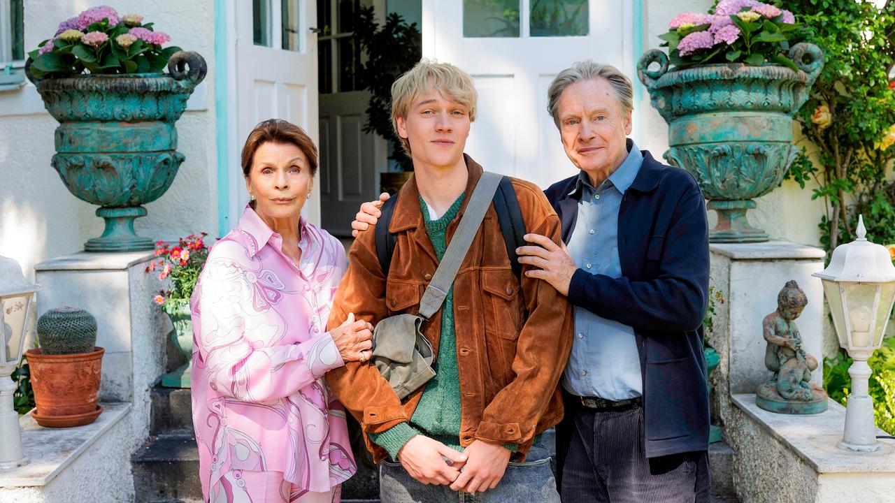 Eine lockige Frau schaut aus der Nähe freundlich zu einem jungen blonden Mann herab.