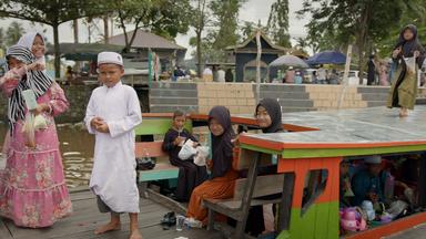 Mehrere Kinder auf einer Barkasse, Mädchen in Kleid und meist mit Kopftuch, Jungen in langem Gewand und mit indonesischer Kopfbedeckung. Im Hintergrund eine Treppe, die zum Wasser führt, sowie mehrere Gebäude, Verkaufsstände und Passanten