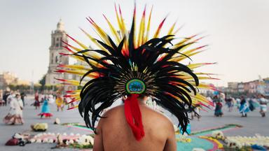 Rückenansicht eines Mannes mit freiem Oberkörper und auffälliger Federkrone mit schwarzen, roten, gelben und weißen Federn, im Hintergrund Menschen in bunter Tracht auf einem Platz unter freiem Himmel.