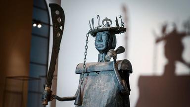 Oberkörper einer Statue aus Metall mit einer Machete in der rechten und einem kleineren Objekt in der linken Hand und einer Kopfbedeckung, an die mehrere unterschiedlich geformte Objekte gesteckt sind