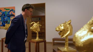 Ein Mann mit Brille betrachtet einen goldenen Tierkopf auf einem Hocker vor ihm, daneben 3 weitere goldene Tierköpfe.