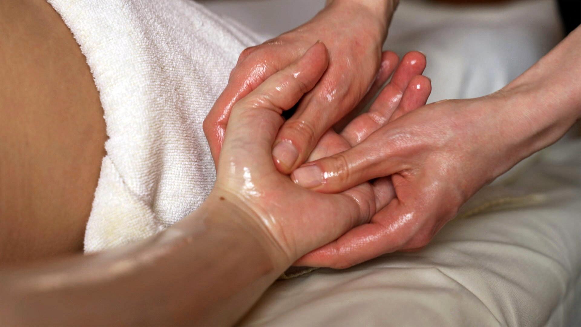 Das Bild zeigt eine Handmassage. Zwei Hände, eine mit Öl bedeckt, drucken auf die andere. Im Hintergrund ist ein Handtuch zu sehen. Die Umgebung wirkt entspannend und gepflegt.