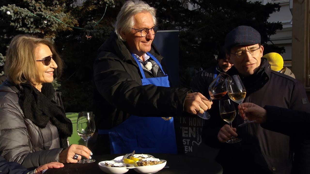 Das Bild zeigt eine Gruppe von vier Personen, die an einem Stehtisch stehen und sich mit Gläsern anstoßen. Im Hintergrund sind Bäume und Weihnachtsdekoration zu sehen, was auf die Vorweihnachtszeit hindeutet. Die Personen tragen wetterangepasste Kleidung; eine Frau hat eine dunkle Jacke und eine schwarze Schal um den Hals, sie lächelt und hat Sonnenbrillen auf. Neben ihr steht ein Mann mit grauen Haaren, der eine blaue Schürze trägt und ebenfalls lächelt. Er wirkt freundlich und gestikuliert mit seinem Glas. Ein weiterer Mann, der dunkle Kleidung trägt und eine Mütze aufhat, steht links von ihm und hält ein Glas in der Hand.   Auf dem Tisch vor den Personen stehen einige kleine Teller mit Snacks, in denen auch Zitrusfrüchte zu sehen sind. Die Szene vermittelt eine festliche Stimmung, und die Personen scheinen sich in einem geselligen Moment zu befinden. Der Kontext des Bildes deutet darauf hin, dass sie an einer wohltätigen Veranstaltung teilnehmen.