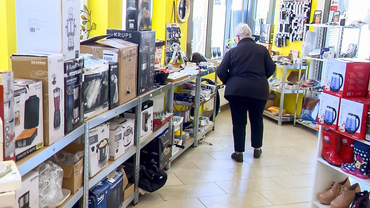 Das Bild zeigt die Innenansicht eines Geschäfts, das eine Vielzahl von Produkten anbietet. Die Wände sind in einem auffälligen Gelb gestrichen, und das Licht kommt durch große Fenster auf der rechten Seite.   In der Mitte des Raums sind Regale aufgestellt, die bis zur Decke gefüllt sind mit Kartons verschiedenster Haushaltsartikel, darunter Küchengeräte und Elektronik. Auf den Regalen liegen viele Produkte in ihren Originalverpackungen, und sie sind chaotisch angeordnet, was darauf hinweist, dass es sich um Retourwaren oder Restposten handeln könnte.   Im Vordergrund ist eine ältere Frau zu sehen, die in einem schwarzen Mantel gekleidet ist und sich etwas abgewandt von der Kamera in Richtung der Regale bewegt. Ihre Haare sind grau, und sie trägt bequeme, dunkle Schuhe. Der Boden besteht aus hellen Fliesen, und die allgemeine Atmosphäre des Geschäfts wirkt lebhaft und einladend.  In der rechten Ecke des Raumes befinden sich zusätzliche Regale mit einer bunten Auswahl an Gummistiefeln und anderen Schuhen, die ebenfalls zum Verkauf angeboten werden.