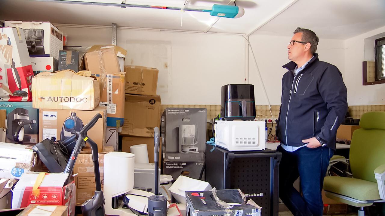 Das Bild zeigt eine Werkstatt, in der zahlreiche Kartons und elektrische Geräte aufbewahrt werden. Im Vordergrund steht ein Mann, der eine dunkle Jacke trägt und in die Werkstatt schaut. Hinter ihm sind viele gestapelte Kartons zu sehen, die teilweise mit Marken wie „Philips“ und „AUTODOC“ gekennzeichnet sind. Unter den Boxen befinden sich verschiedene Haushaltsgeräte, darunter eine Küchenmaschine und ein Staubsauger. Die Werkstatt wirkt unordentlich, mit einem vollen Raum, der wenig Platz für Bewegungen lässt. Im Hintergrund sind graue Wände und ein Fenster mit einem Gitter sichtbar. Allerdings gibt es auch ein paar verdeckte Bereiche, die nicht klar einsehbar sind. Insgesamt vermittelt das Bild den Eindruck eines Raumes, der mit Retourwaren und zum Verkauf gedachten Produkten gefüllt ist.