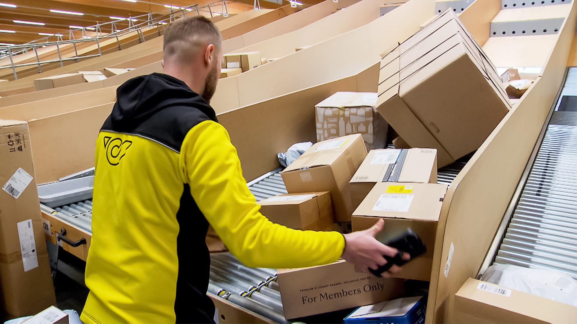Das Bild zeigt einen Mitarbeiter in einem Paketverteilzentrum. Er trägt eine schwarz-gelbe Arbeitsjacke und ist dabei, Kartons zu sortieren. Die Umgebung ist geprägt von lange gestreckten Förderbändern, auf denen zahlreiche Päckchen und Pakete liegen. Die Pakete sind in verschiedenen Größen und Formen, einige sind beschriftet, während andere versiegelt sind. Im Hintergrund sind weitere Reihen von Paketen sichtbar. Die Decke des Zentrums ist aus Holz und es gibt eine helle Beleuchtung. Der Mitarbeiter ist von der Seite zu sehen und scheint in konzentrierter Arbeit zu sein, während er ein Paket in der Hand hält.