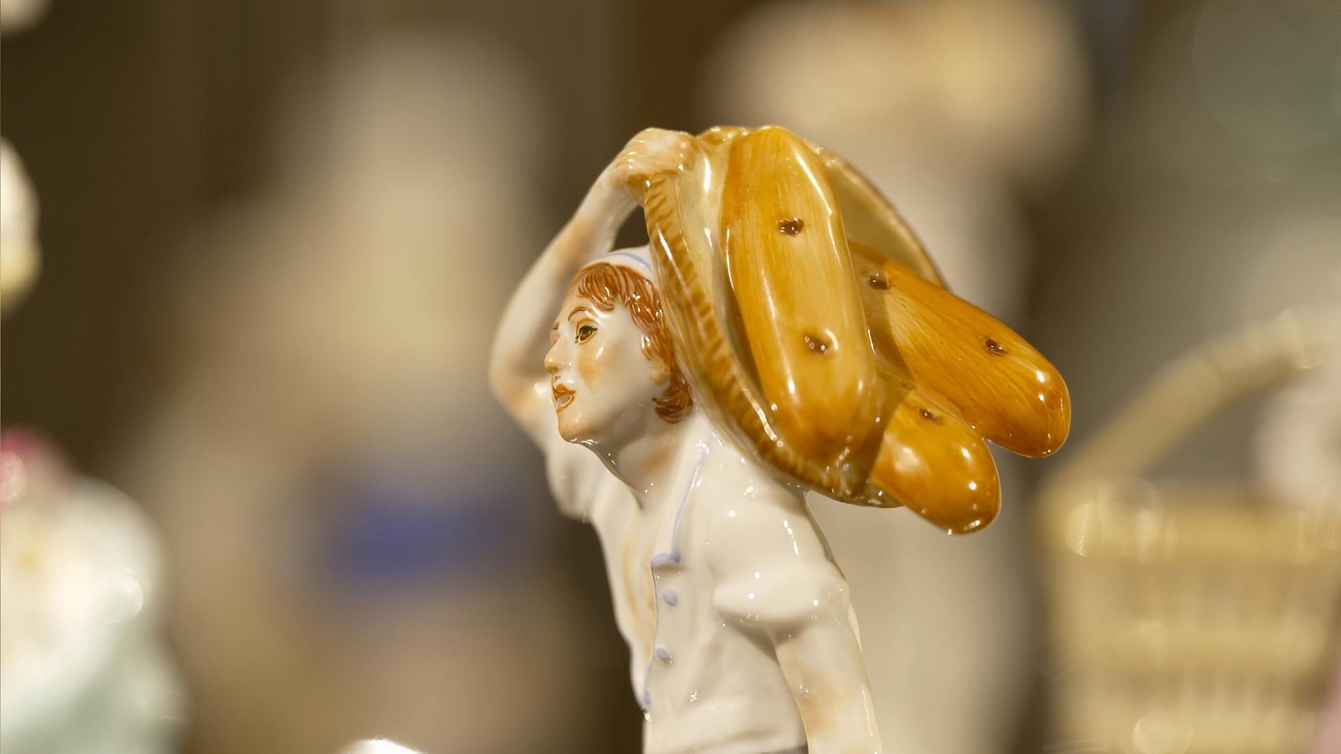 Nahaufnahme einer bemalten Meissener Porzellanfigur eines Brotträgers mit großer Brezelform auf der Schulter, im unscharfen Museumsambiente.