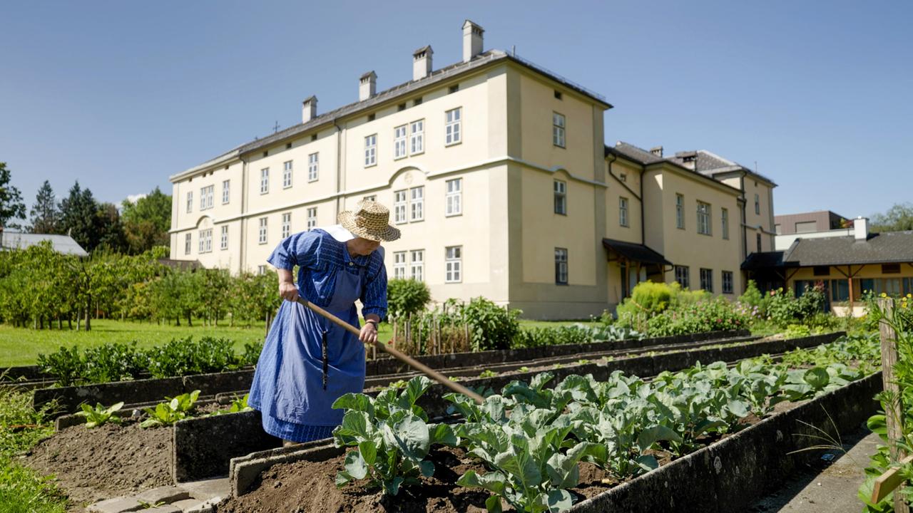Das Bild zeigt eine Frau, die im Garten eines Klosters arbeitet. Sie trägt ein blaues, traditionelles Kleid und einen Strohhut. Die Frau ist dabei, den Boden mit einer Hacke zu bearbeiten, um Gemüse anzupflanzen. Der Garten ist in mehrere Beete unterteilt, auf denen vorwiegend Kohlpflanzen sichtbar sind.   Im Hintergrund steht ein großes, gelbes Gebäude, das wahrscheinlich das Kloster ist. Es hat mehrere Fenster und ein einfaches, aber elegantes Design. Um das Gebäude herum sind verschiedene Pflanzen und Sträucher angelegt, die dem Ort ein angenehmes und gepflegtes Aussehen verleihen. Der Himmel ist klar und blau, und es ist sonnig.   Die Gesamtstimmung des Bildes wirkt friedlich und ländlich, und es vermittelt ein Gefühl von Arbeit im Freien.