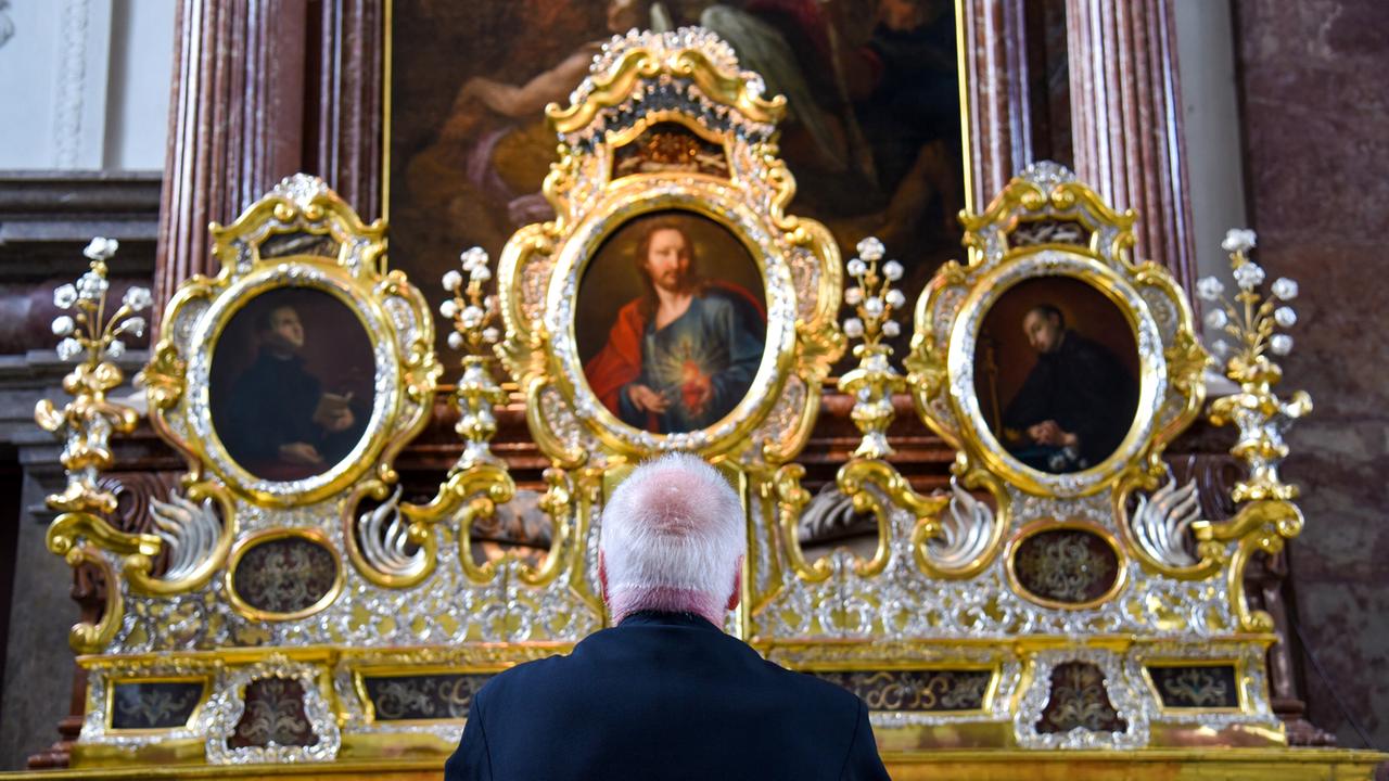Das Bild zeigt einen älteren Mann, der in einem kirchlichen Raum vor einem prächtigen Altar steht. Der Mann hat graue Haare und trägt einen dunklen Anzug. Sein Blick ist zum Altar gerichtet. Der Altar ist aufwendig gestaltet, mit goldenen und silbernen Verzierungen. In der Mitte des Altars befindet sich ein großes Gemälde, das Jesus darstellt, umgeben von zwei weiteren kleineren Gemälden, die wahrscheinlich Heilige oder Jesuiten zeigen. Der Hintergrund des Altars ist in dunkleren Farben gehalten, während die Verzierungen hell leuchten. Die Atmosphäre wirkt andächtig und feierlich, passend zu einem religiösen Kontext.