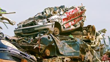 Das Bild zeigt einen Schrottplatz, auf dem zahlreiche alte, beschädigte Autos übereinander gestapelt sind. Es sind verschiedene Fahrzeugmodelle sichtbar, die stark verrostet und verformt sind. Ein Auto in der Mitte trägt rote Schrift mit dem Text "Dominique Michelle in Love." Der Hintergrund ist meist wolkenverhangen und wirkt trüb. Der Anblick ist geprägt von einer chaotischen Ansammlung von Metall und teilweise auch anderen Materialien, die auf eine mangelhafte Abfallwirtschaft hinweisen. Der Kontext des Bildes behandelt die Problematik der Müllentsorgung und das Thema einer "Gesellschaft ohne Müll."