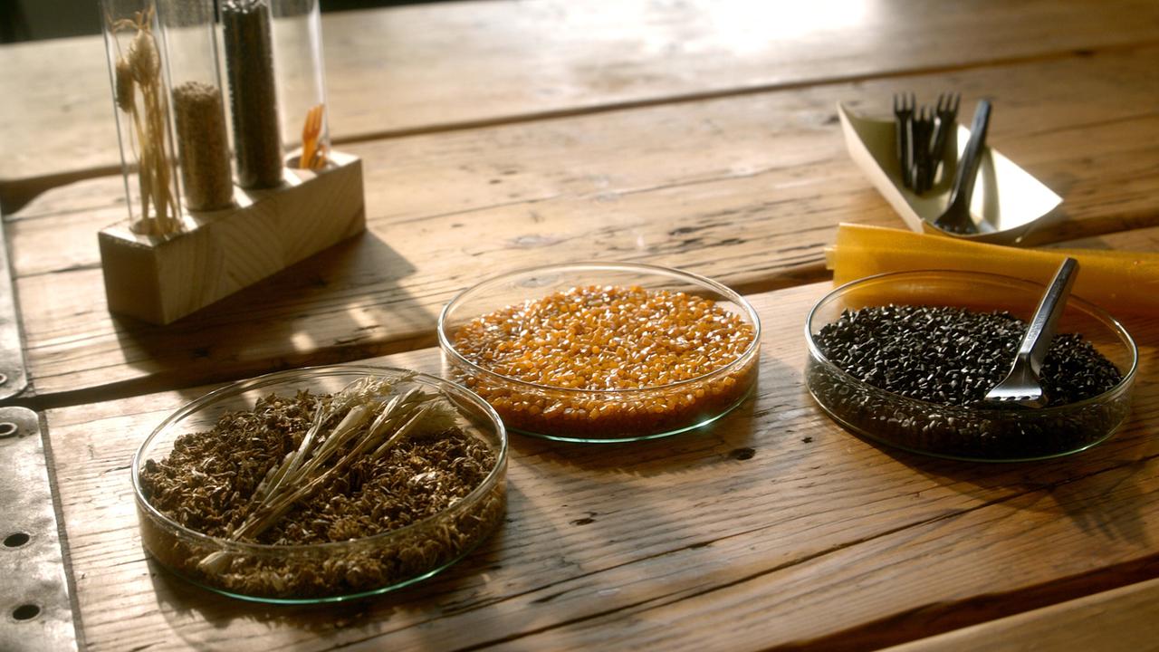 Das Bild zeigt eine angeordneten Zusammenstellung von verschiedenen Materialien auf einer Holzoberfläche. In der Mitte sind drei klare Gläser mit unterschiedlichen Inhalten.   - In einem Glas befindet sich eine braune, faserige Substanz oder Samen. - Ein weiteres Glas enthält orangefarbene, kleine Körner oder Granulate. - Das dritte Glas enthält schwarze, kleine Partikel oder Körner.  Neben den Gläsern sind mehrere durchsichtige Röhrchen auf einem Holzblock platziert, die mit verschiedenen Zutaten gefüllt sind. Im Hintergrund sind teilweise schwarze, plastikähnliche Besteckteile zu sehen, die ordentlich in einem weißen, geschwungenen Behälter liegen. Es sind auch einige durchsichtige, gelbe Folien oder Materialien sichtbar.  Die Holzoberfläche weist eine natürliche Maserung auf, und die gesamte Anordnung wirkt sehr strukturiert und sorgfältig arrangiert, passend zum Thema nachhaltige Materialien und Recycling.