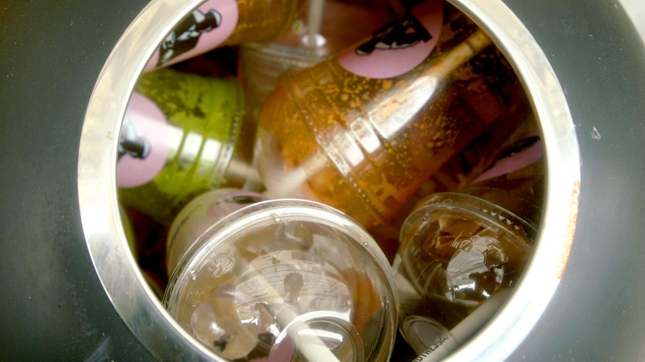 Das Bild zeigt eine Ansammlung von Plastikmüll, die in einem Behälter liegt. Zu sehen sind verschiedene Arten von Plastikbechern, darunter transparente Becher mit Deckeln und bunten Strohhalmen. Einige Becher sind grün und braun, während andere klar sind, und alle scheinen mit Flüssigkeitsresten gefüllt zu sein. Die Becher sind übereinander gestapelt, was darauf hindeutet, dass sie nicht ordnungsgemäß entsorgt wurden. Der Behälter hat einen metallischen Rand und ist insgesamt dunkel, was einen Kontrast zu den bunteren Bechern bildet. Das Bild thematisiert die Problematik der Abfallwirtschaft und den Umgang mit Plastikmüll.