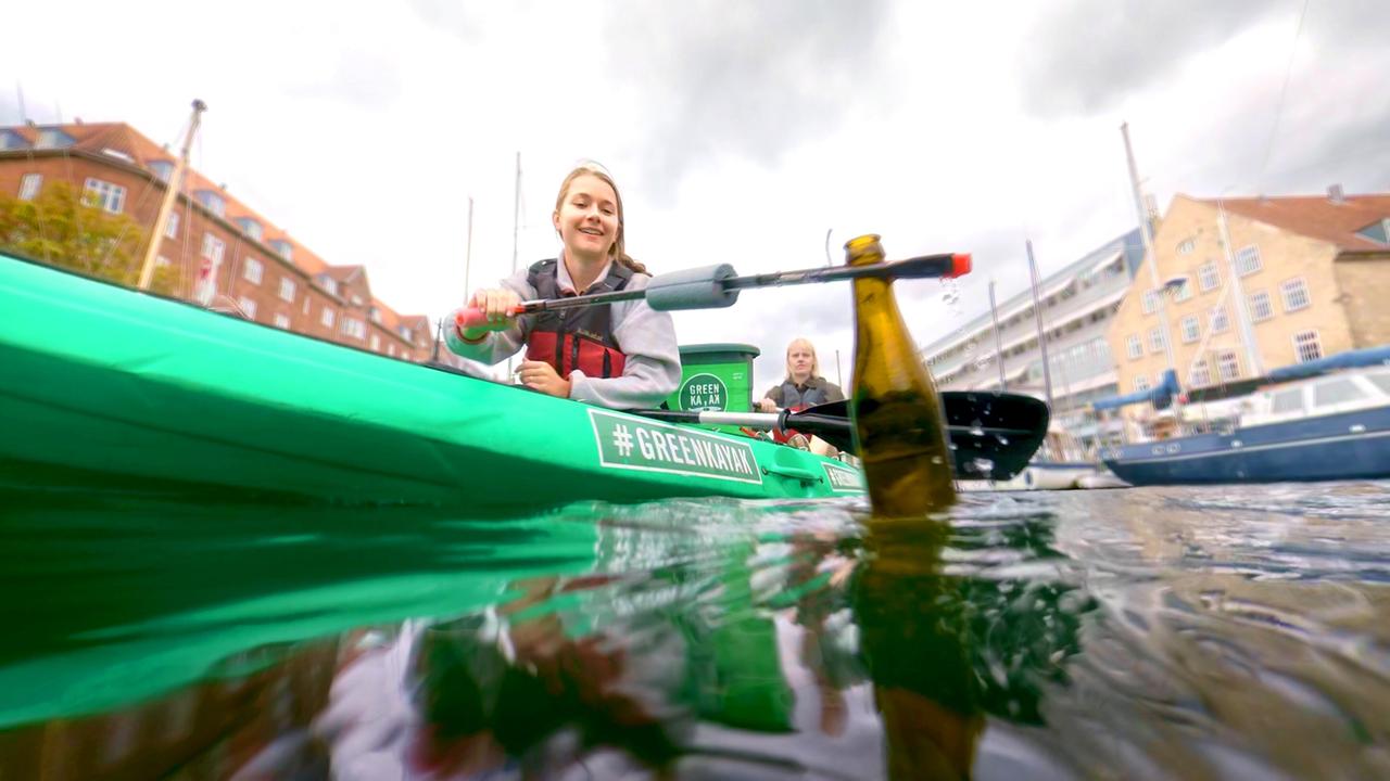 Das Bild zeigt eine Szene aus dem Projekt „Green Kayak“, das sich mit dem Einsammeln von Müll in Gewässern beschäftigt. Im Vordergrund ist ein grünes Kajak zu sehen, in dem zwei Personen paddeln. Eine der Personen lächelt und hält einen Paddel in der Hand, mit dem sie ein Stück Müll, eine Flasche, aus dem Wasser fischt. Die andere Person sitzt hinter ihr im Kajak.   Der Hintergrund zeigt eine städtische Umgebung in Kopenhagen, mit mehrstöckigen, backsteinartigen Gebäuden, die von Bäumen umgeben sind. Am Ufer sind auch einige Boote zu sehen. Der Himmel ist bewölkt, was auf eine mäßige Wetterlage hindeutet. Das Wasser im Kanal wirkt klar, und die Aktion vermittelt den Eindruck eines umweltbewussten Engagements.   Das Bild ist Teil eines Films, der die Auswirkungen von Abfall in der Umwelt thematisiert und Initiativen zur Bekämpfung des Müllproblems beleuchtet.