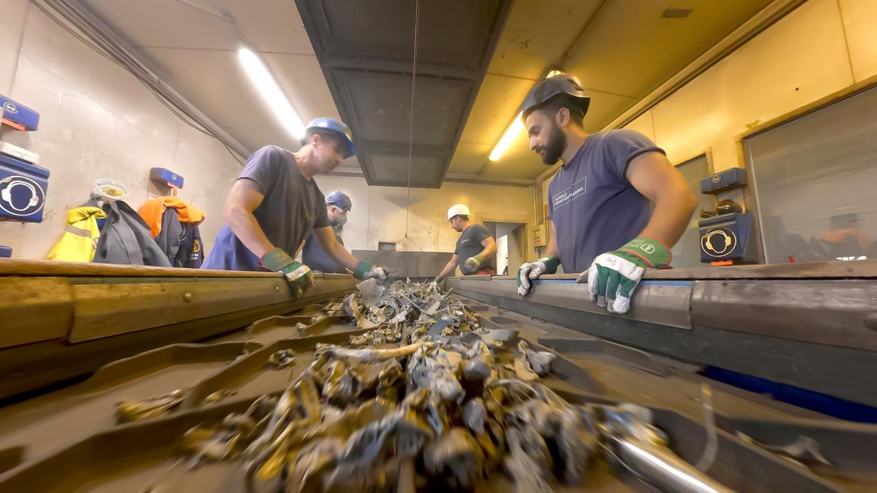 Das Bild zeigt eine Innenansicht einer Recycling- oder Sortieranlage. In einem hell erleuchteten Raum sind mehrere Personen mit Schutzhelmen und Handschuhen am Arbeiten. Sie stehen um einen Förderband, auf dem Abfallmaterialien liegen, die sortiert werden. Die Abfallstücke sind überwiegend aus Metall, darunter zerknüllte und verformte Teile, die eine Vielzahl an Formen und Größen aufweisen.  Die Arbeiter sind verschiedene Gesichtsausdrücke wie Konzentration und Engagement zu erkennen, während sie ihr Augenmerk auf die Materialien richten. Im Hintergrund sind Wände aus Beton und einige Geräte oder Werkzeuge sichtbar, die im Recyclingprozess verwendet werden. Die Beleuchtung des Raumes ist hell, um die Sicht zu verbessern. Es herrscht eine geschäftige, aber ordentliche Atmosphäre, die auf die Wichtigkeit der Abfallwirtschaft hinweist.